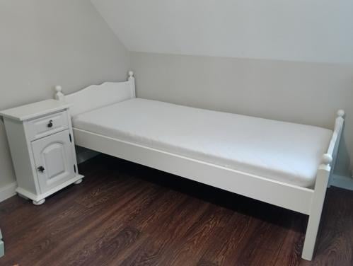 Wit eenpersoons bed met lattenbodem en nachtkastje.