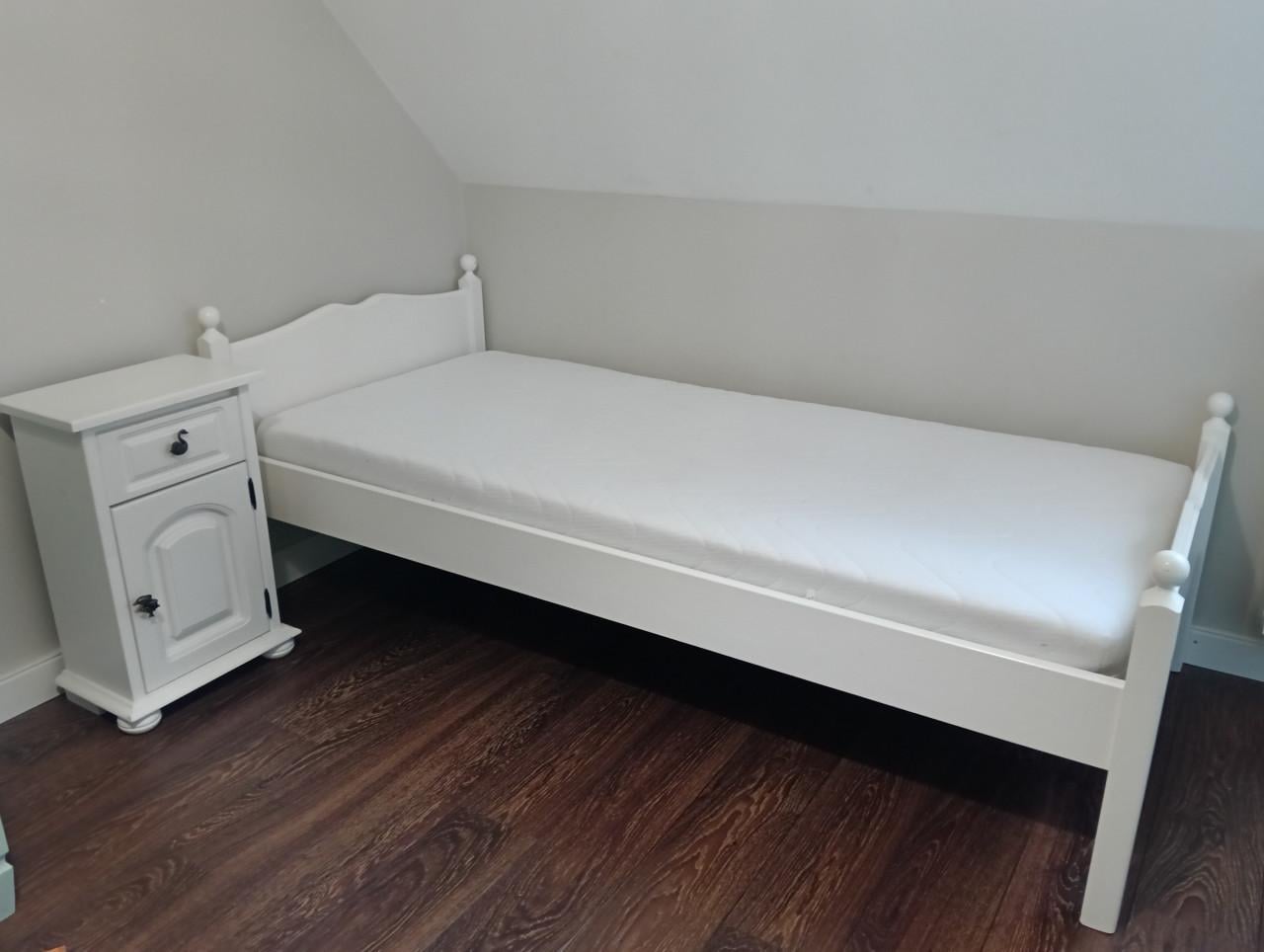 Wit eenpersoons bed met lattenbodem en nachtkastje.
